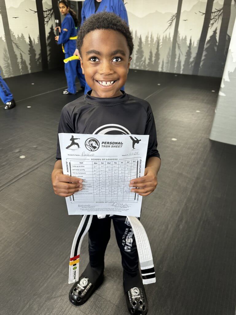 a young boy, in martial arts uniform, holding a worksheet and smiling.